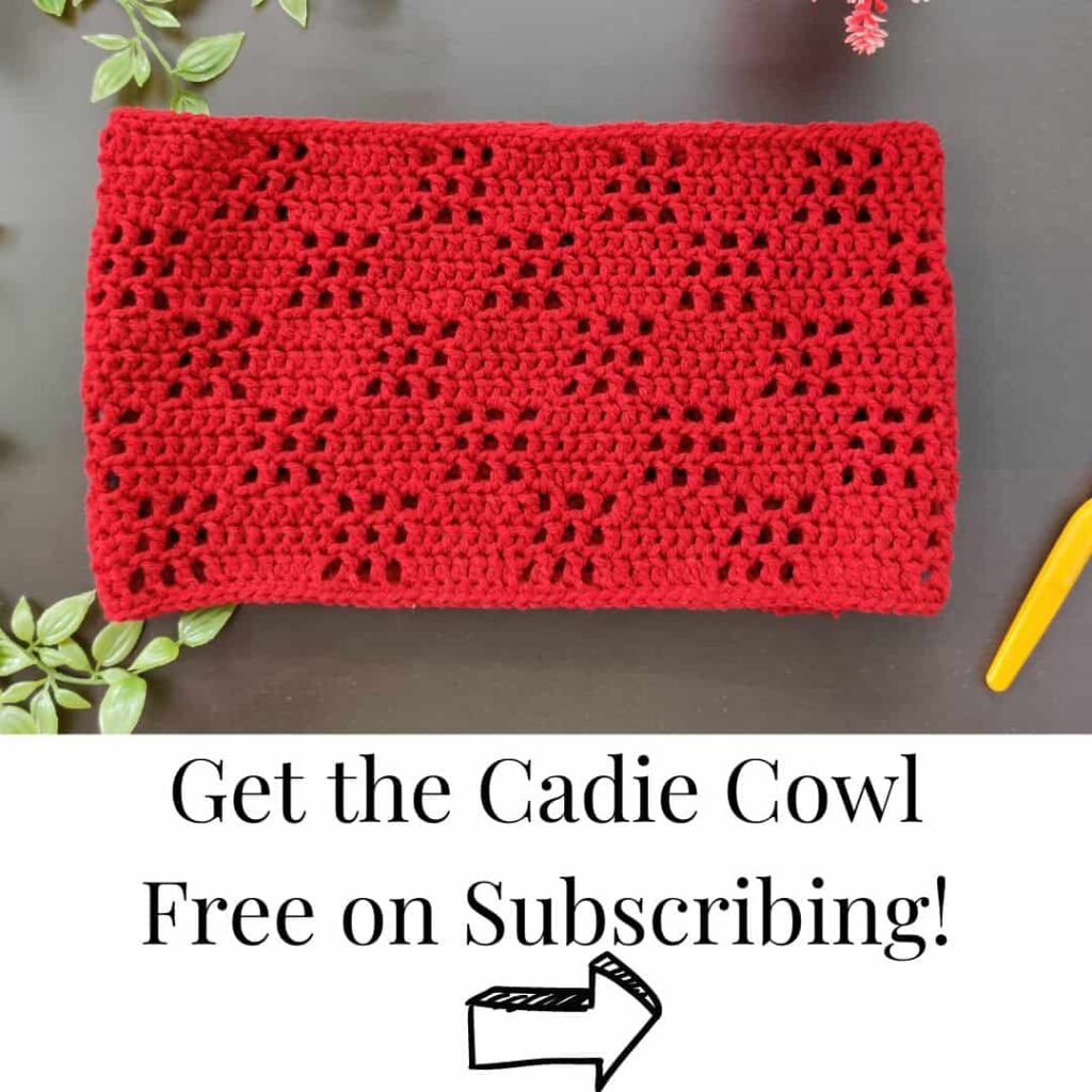 cadie cowl in red lying against a blackish table with leaves and a crochet hook with text get the cadie cowl free on subscribing